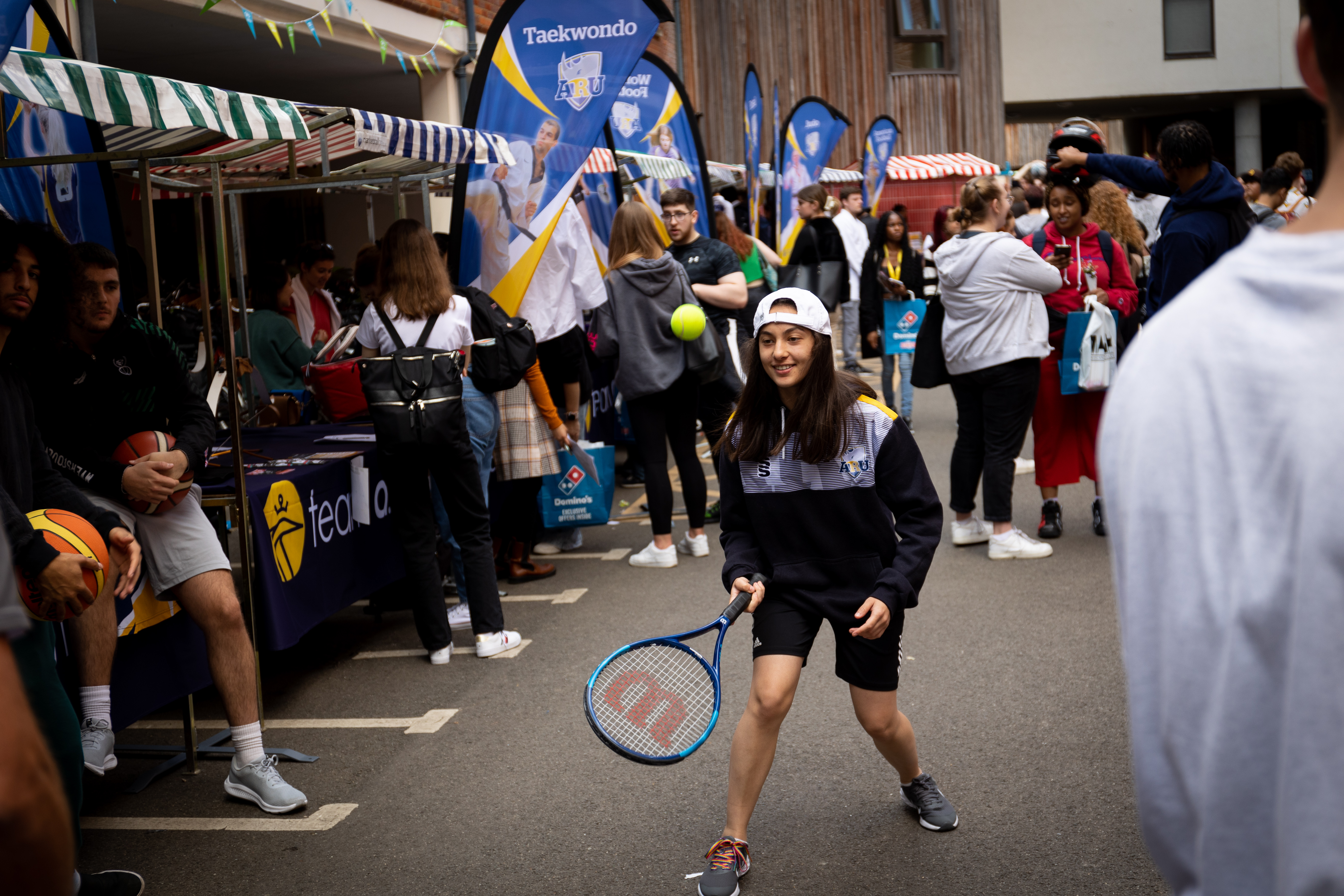 Woman hitting a ball with a tennis racket surrounded by Welcome Fair stalls